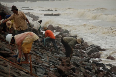 Arbeiders leggen basaltblokken, de zee is woelig en slaat er bijna overheen